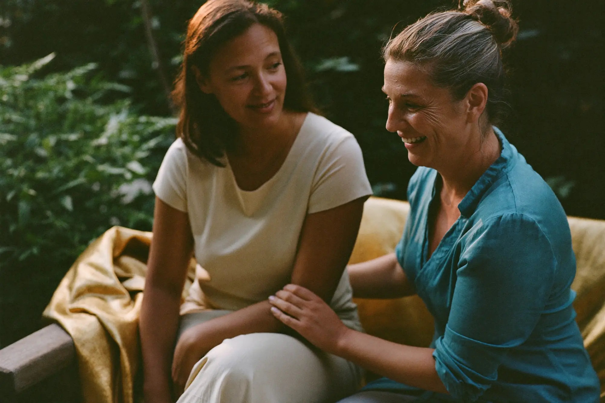Two women sitting on a garden bench in evening light, one laughing mid-conversation.