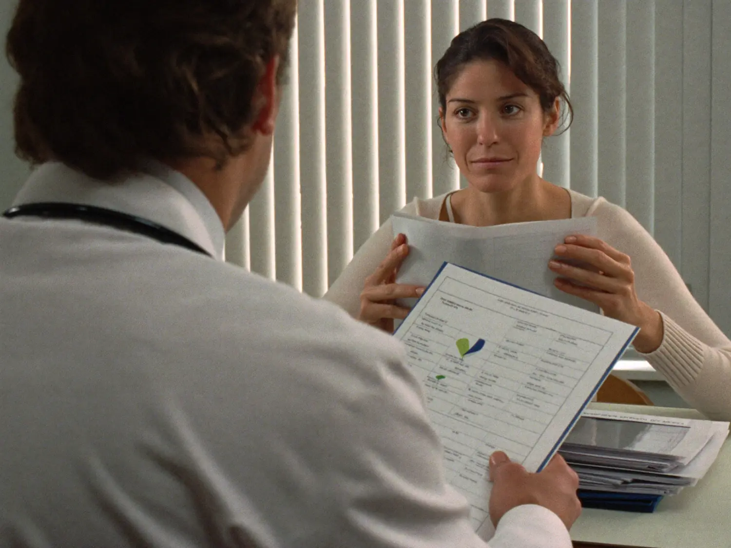 A woman in a doctor's office holding a sheet of test results, looking across the desk at the doctor.