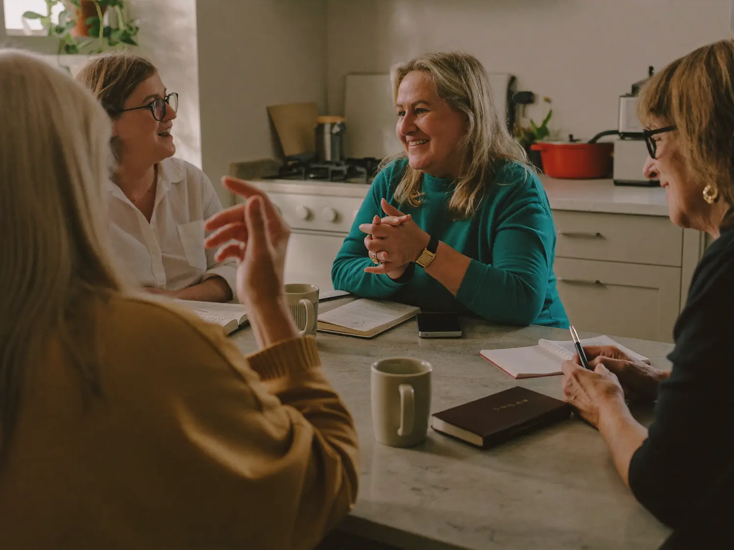Four women talking around a kitchen table, with open notebooks and mugs of coffee between them.