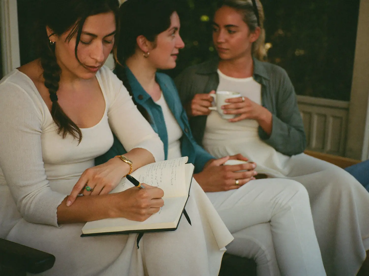 Three women sitting together on a porch, one writing in a journal while the others talk over coffee.
