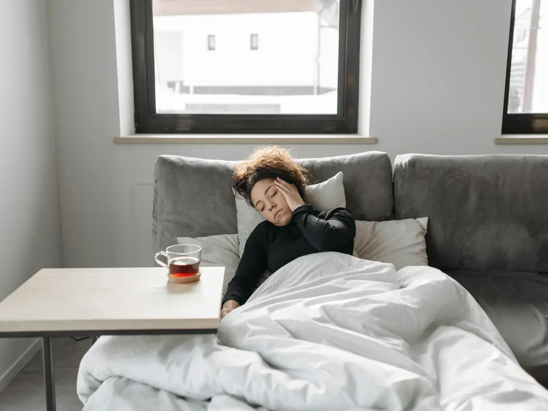 A woman resting on a sofa under a duvet, a cup of tea on the table beside her.