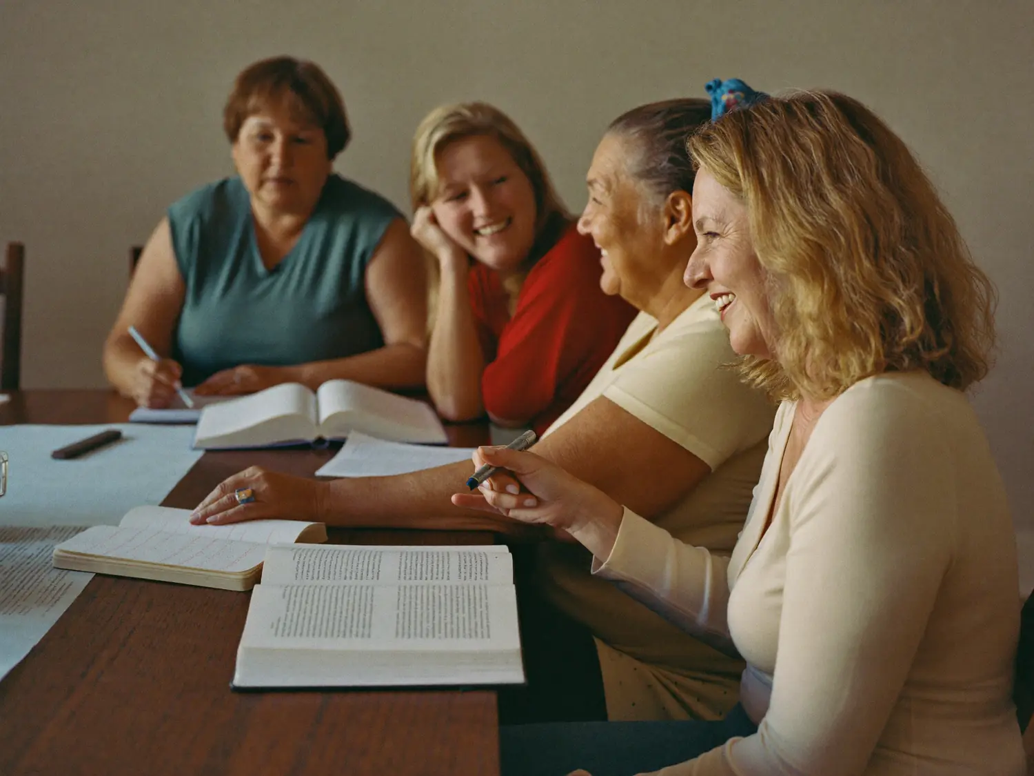 Four women sharing a laugh around a table of open books and notebooks.