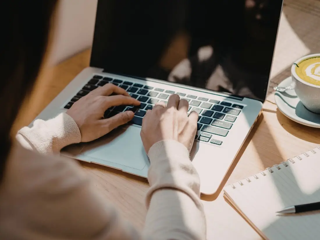 Hands typing on a laptop at a wooden table beside a notebook and a cup of coffee.